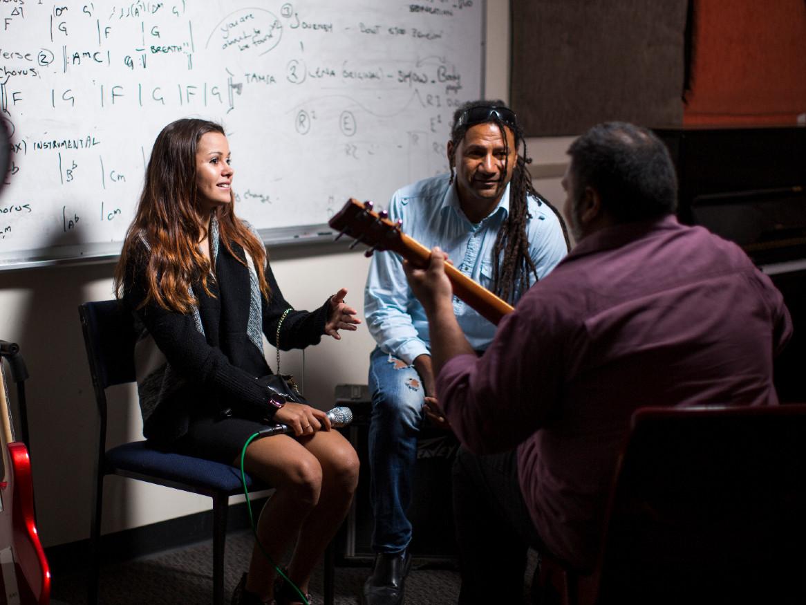 a girl holding a microphone sits with a man facing a second man playing a guitar in a classroom