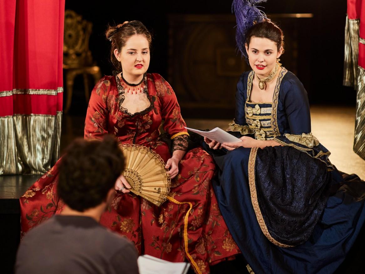 two female music theatre students sit on stage in costume speaking with a staff member