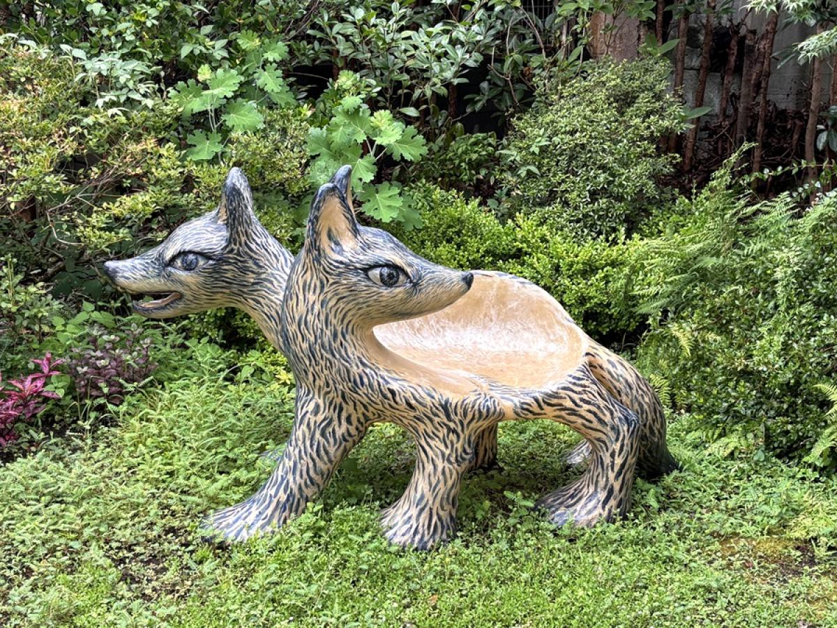 A bench in the shape of a two-headed wolf is outdoors surrounded by greenery