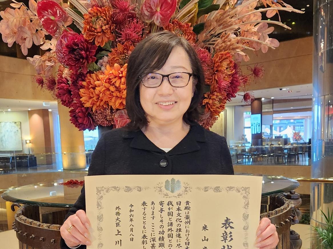 Shoko Yoneyama stands and smiles, holding her Japanese Foreign Minister’s Commendation award